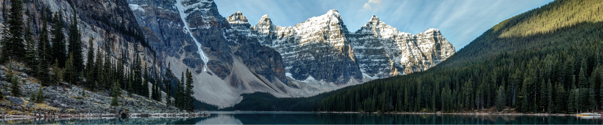 Blue stream of water surrounded by green forestry on the right side and the bottom of a rocky grey mountain on the left side